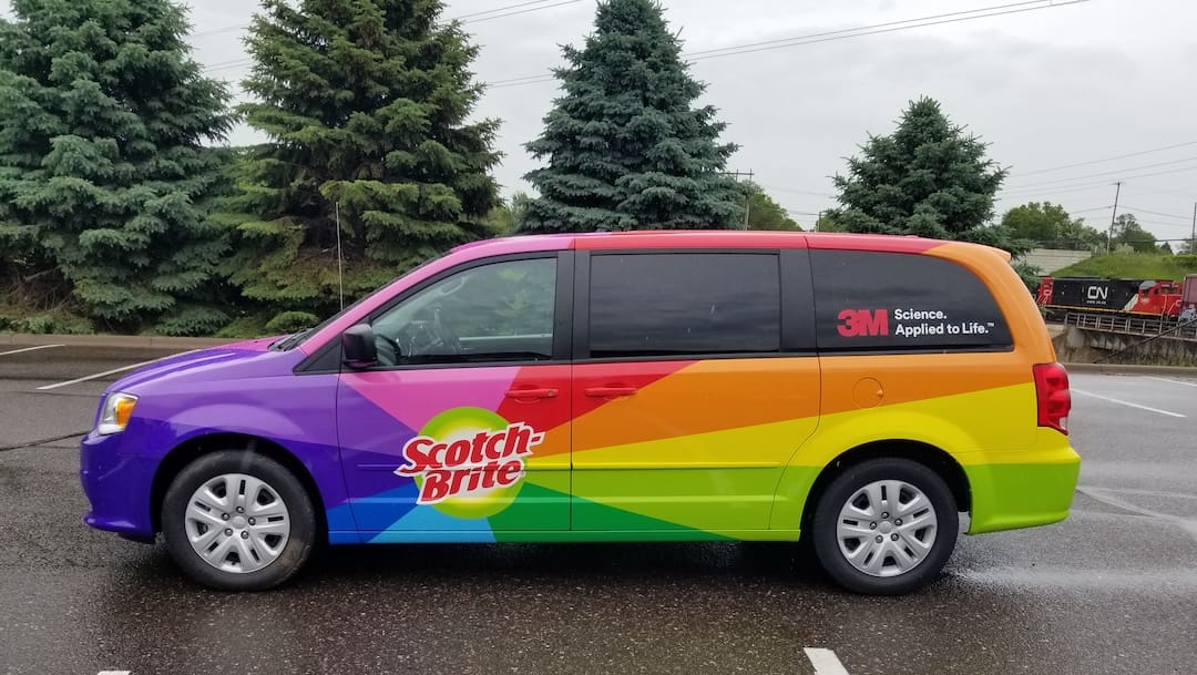 Brand Ink wrapped a vehicle for 3M Scotch Brite in rainbow colors. The vehicle sits outside in a parking lot in front of evergreen trees.