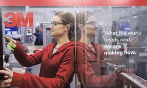 A 3M employee in a red hoodie operating factory equipment, reflected in a glass panel next to the slogan "What the world needs next, we are making now."