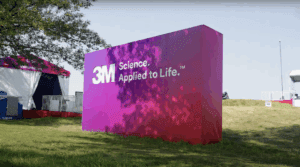 A purple wall stands in the middle of a grassy area in front of a tent. The wall reads "3M Science Applied to Life."