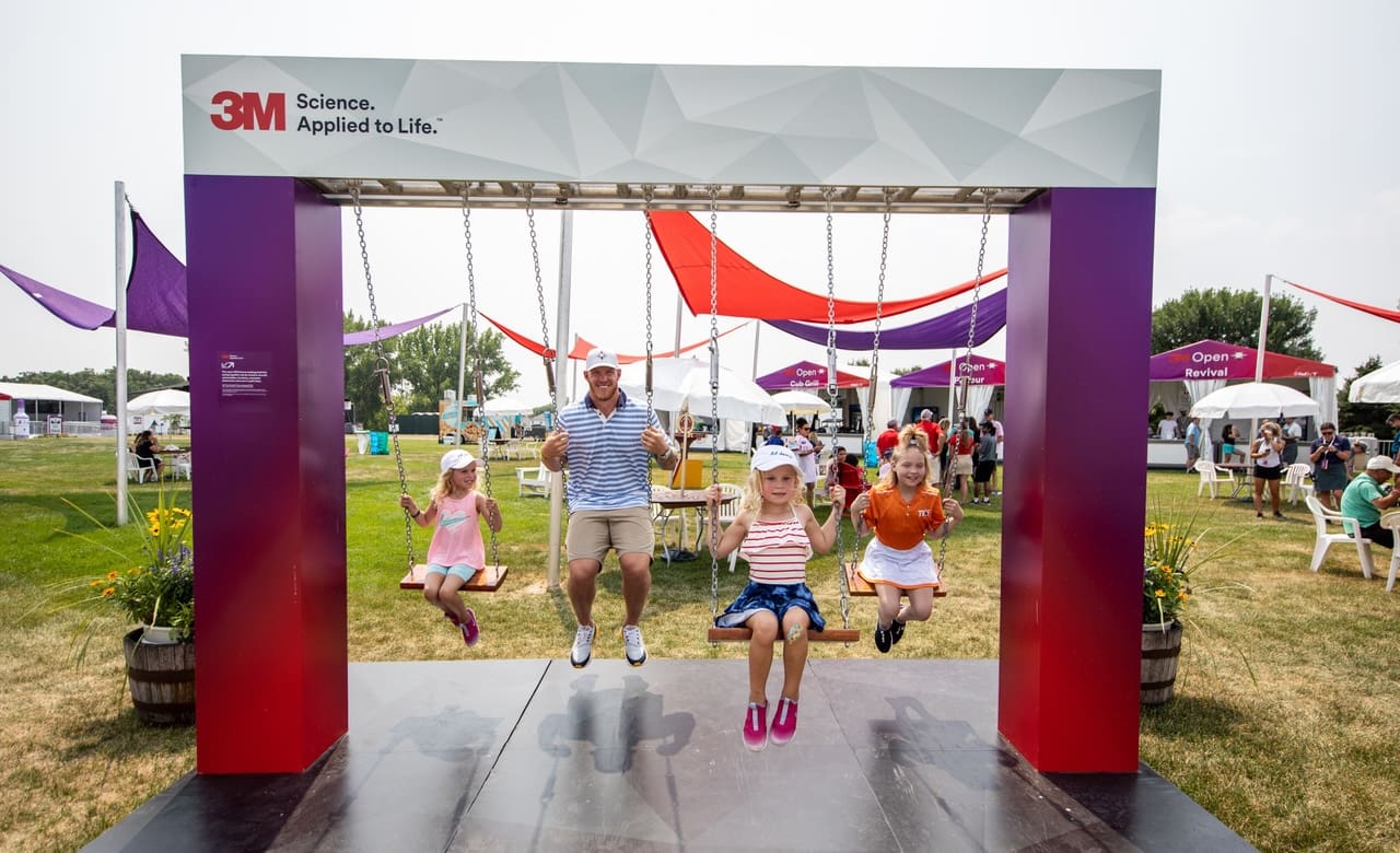 three children and their father swing on a swing set at the 3M open in Minneapolis, MN. The swing set is branded 3M with printed graphics installed by Brand Ink.