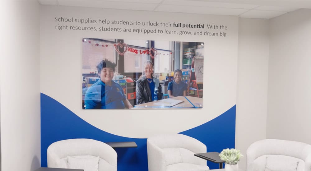 Brand ink installed a mounted printed acrylic on a wall in Kids In Need Foundation's headquarters: It is a picture of three students sitting at a table in a classroom. Over the image is a vinyl decal that reads "School supplies help students unlock their full potential. With the right resources, students are equipped to learn, grow, and dream big."