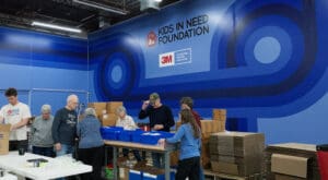 Several volunteers sort through school supplies in front of a large, saturated blue wall with designs, the Kids in Need Foundation logo, and 3M's logo next to a small statement that reads, "Proud to help bring this space to life."