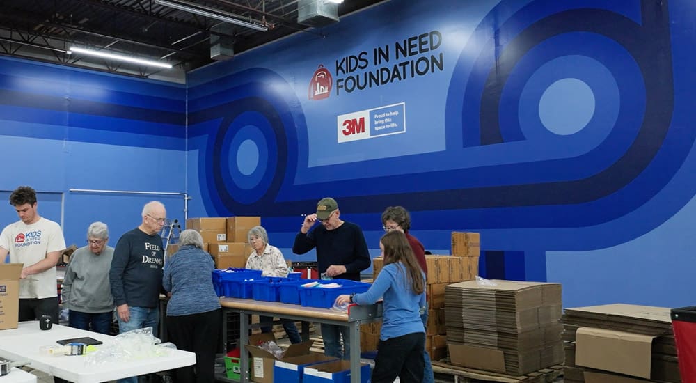 Several volunteers sort through school supplies in front of a large, saturated blue wall with designs, the Kids in Need Foundation logo, and 3M's logo next to a small statement that reads, 