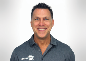 Smiling man in a gray polo shirt with a 'brand ink' logo on the chest, facing the camera.